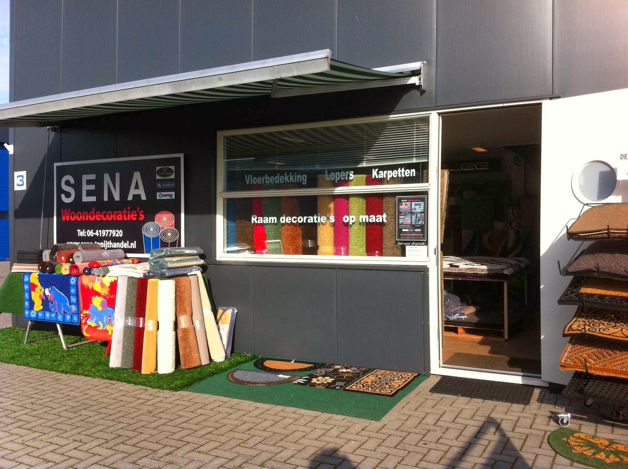 De gevel van SENA Woondecoratie's aan de Lange Baan in Steenwijk, met karpetten en tapijtrollen voor de deur
